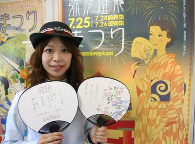 弥彦燈籠まつりのポスターを背に、そのイラストと同じにデザインしたうちわを持つ弥彦観光協会・小川泰枝弥彦観光駅長