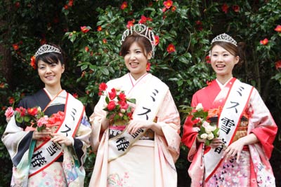 ミス雪椿3人が加茂市民体育館前のユキツバキの花をバックにスチール写真を撮影、干だらからミス雪椿の滝沢さん、ミス雪椿クイーンの吉崎さん、ミス雪椿の徳橋さん。