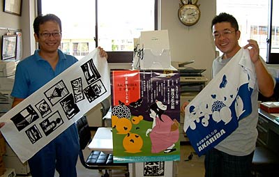 スタンプラリーでプレゼントするオリジナル手ぬぐいを手に来場を呼びかける左から柳生田副委員長、佐竹委員長