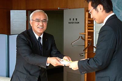 香典返しとして三条市に寄付する100万円を国定市長(右)に手渡す故吉田盛之助さんの長男、英達さん(左)