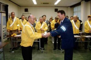 燕市交通安全協会で行われた「高齢者ピカッと作戦」出発式でを行い、羽豆交通課長から登坂会長に反射材ベルトとたすきを手渡す