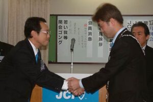 加茂青年会議所の牛腸理事長(左)からプレジデンシャルリーフと理事長バッジを引き継ぎ、握手する桑原次年度理事長(右)