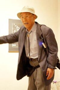 ことし6月6日に燕市産業史料館で開かれた捧武写真展の作品解説会のときの捧さん、肩からカメラを下げて開設する