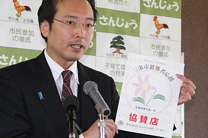 定例記者会見で避難者応援サービス「ひめさゆりカード」の協賛店に152店の登録があったことを紹介、協賛店に掲示されるヒメサユリマークの入った張り紙を示す国定市長