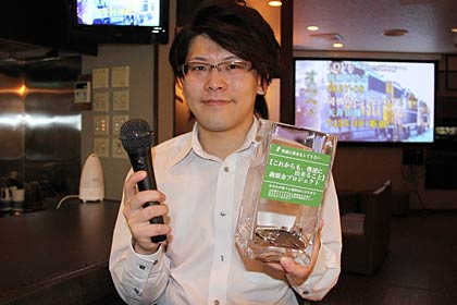 カラオケスナック亀亀の結城店長、右手にカラオケマイク、左手に義援金