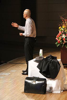 帽子を革のかばん、ジャケットをテーブルに置き、ピンマイクで熱く話す菊地弁護士