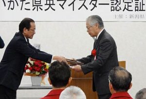 26日開かれた第6回にいがた県央マイスター認定証授与式で岡田県三条地域振興好局長(左)から認定証をうけうるきせる職人の飯塚さん(右)