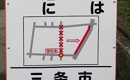  工事場所の看板に描かれた切り替えの図説、赤い「×」のあるのが閉鎖区間、赤い実線で描かれた部分が切り替えになった自転車歩行者道、ちょうど赤い「○」が描かれた所にリースを掲げた