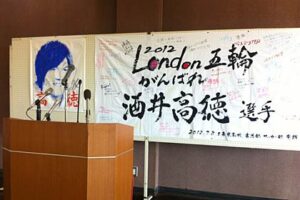 10日の国定三条市長の記者会見で国定市長の背中を飾る高徳選手応援の寄せ書きや高徳選手の顔が描かれたゲーフラ