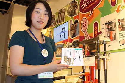 燕市産業史料館がスプーン製作の疑似体験で作ったミニスプーンに取り付けるストラップとチェーンの販売を開始、ストラップとチェーンを持つ職員の後ろがスプーン製作の疑似体験コーナー