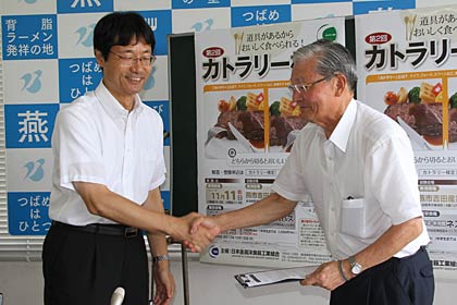 記者会見で受検申込票を記入した鈴木市長と握手する田中さんｎ