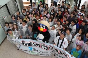 23日、燕市の農業体験田「スワローズ・ライスファーム」で行われた稲刈りでつば九郎を日帰りバスツアーの参加者が囲んで記念撮影