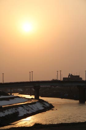 6日夕方、三条市林町地内の五十嵐川右岸から下流側に浮かぶ夕日はかすで輪郭がぼやけて見えた