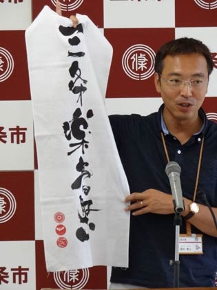 「被災地応援手拭いプロジェクト」で製作したオリジナル手ぬぐいを記者会見で紹介する国定三条市長