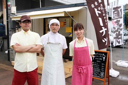 佐久間食品が同社敷地にオープンした「町のドーナツ屋さん」の前で二代目佐久間社長（中央）と三代目康之さん夫婦