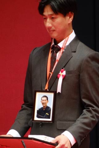 三条市島田1、スポーツサイクルショップ経営の坂本敏明さんの遺影を首から下げて代わりに表彰状を受け取った長男の聡さん