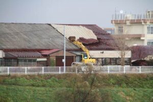 屋根の左側のトタンがはがれ、残った右側部分にパワーショベルのアームを置いてトタンがはがれないように対応