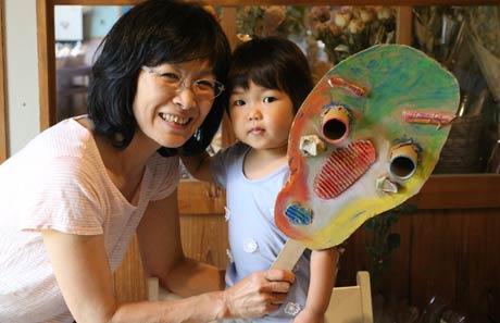 三条おやこ劇場理事長の兼古さんと1歳の孫娘、手にしているのはワークショップで作る空想楽器、ムラドクスマ