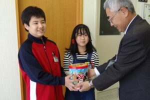 三条市社会福祉協議会の小林事務局長に缶に詰まった6万円余りの善意を手渡す月岡小のスマイルライフ委員会委員長の6年生高浪君とJRCエコ委員会委員長の6年生岡田さん