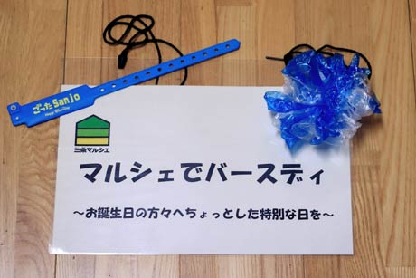 「マルシェ de バースデー」で5月生まれにプレゼントされる特製リストバンドと5月生まれの人が特典を受けられることを示す店の名札に取り付ける青い造花