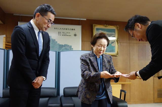 国定市長(右)に2億円の寄付の目録を手渡す捧ミヨエさん(中央)と捧雄一郎社長(左)