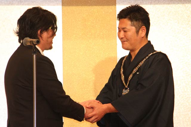 本間前理事長(左)から理事長バッジを受け継いで握手する神田理事長