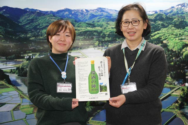 天野尚撮影の下田地区の棚田の写真をバックに「環酒の陣」のちらしを持つ市の担当者