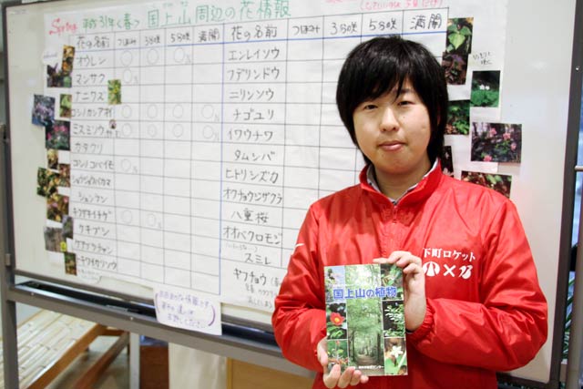 「道の駅 国上」に掲示している国上山周辺の山野草の開花状況と『西蒲原の自然ガイドブックNo.13　国上山の植物』を手に担当の地域おこし協力隊の岡田さん