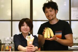バナナ&チョコレートのクレープを手にTREEのメニュー開発担当の星田さん(左)とそれに使ったバナナを手にカネギフルーツの信賀店長