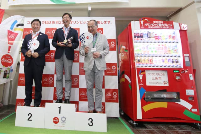 三条市役所に設置された聖火リレーメモリアル自動販売機とフォトブース、左からコカ・コーラボトラーズジャパン新潟支社三条支店野坂上支店長、国定市長、若山副市長