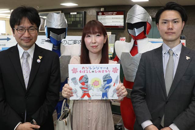 絵本『カジレンジャーとまぼろしのひめさゆり』と左から三条商工会議所青年部の山村会長、絵と文のあだちさん、DX広報委員会の佐藤委員長、後ろに燕三条戦隊スーパーカジレンジャー