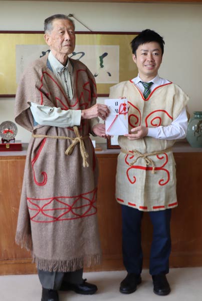 貫頭衣を着て高橋さん(左)から滝沢市長に目録を手渡す
