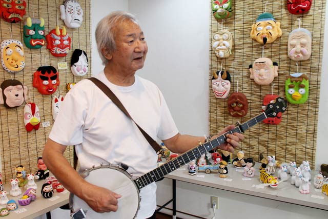 自身の張り子作品に囲まれてネックが長いバンジョーを弾いて歌う古川豪さん